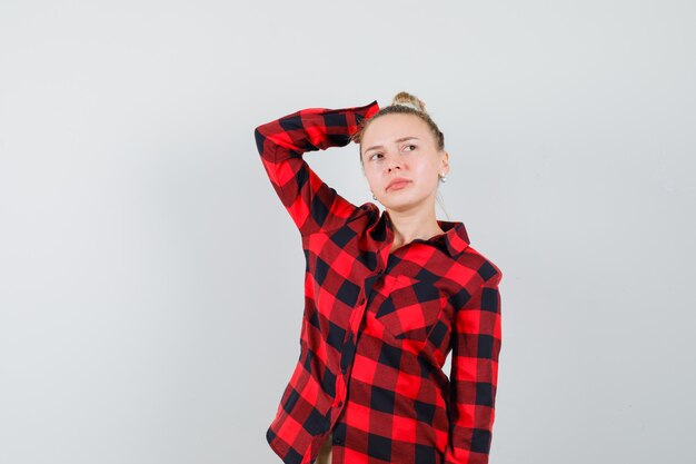 Young woman keeping raised hand on head in checked shirt and looking hesitant