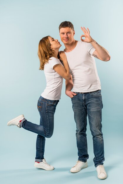 Young woman kissing her smiling boyfriend showing ok sign against blue backdrop