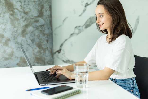 Young woman in kitchen eating berries and using laptop. Happy female looking at laptop while standing in kitchen in morning.