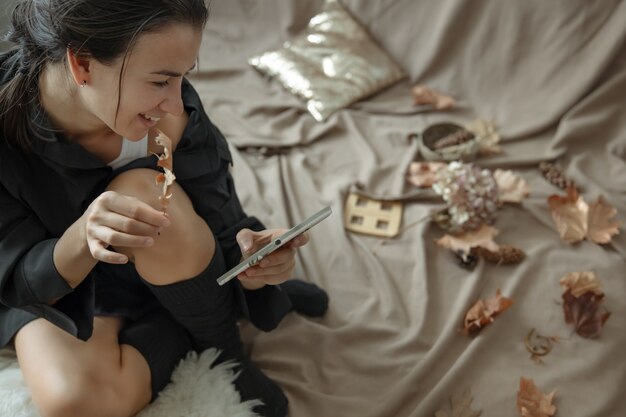 A young woman in knitted stockings uses the phone in a cozy bed, among the autumn leaves.