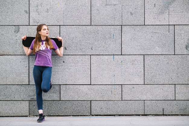 Young woman leaning against a grey tiled urban wall standing on one leg