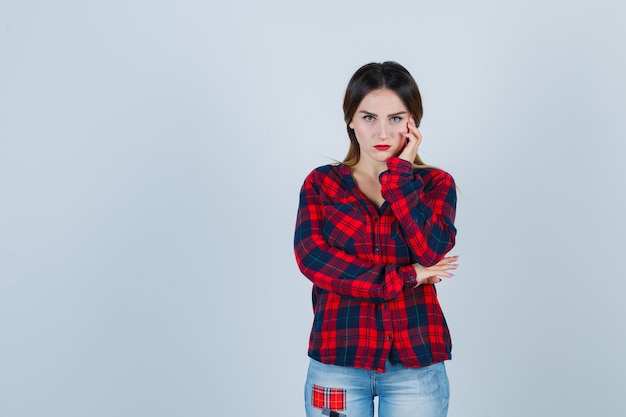 Young woman leaning cheek on hand in checked shirt, jeans and looking upset , front view.