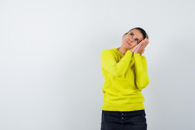 Young woman leaning cheek on hands in yellow sweater and black pants and looking happy 