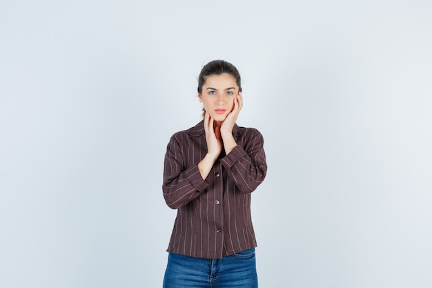 Young woman leaning cheek on palm, with hand on jowl in striped shirt, jeans and looking charming , front view.