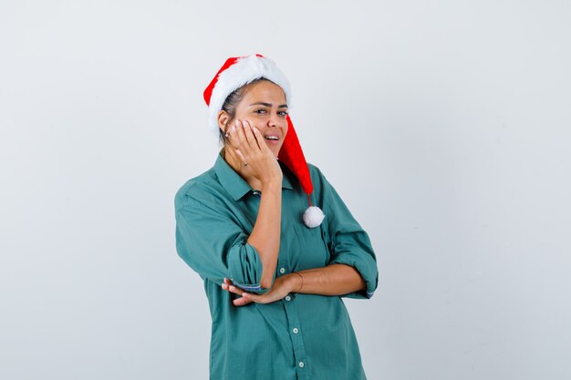 Giovane donna che si appoggia guancia sul palmo alzato in camicia, cappello da babbo natale e si vergogna. vista frontale.