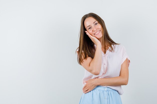 Молодая женщина, опираясь щекой на поднятую ладонь в футболке, юбке и выглядела веселой, вид спереди.