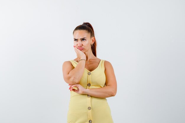 Young woman leaning on chin with palm in yellow dress and looking upset , front view.