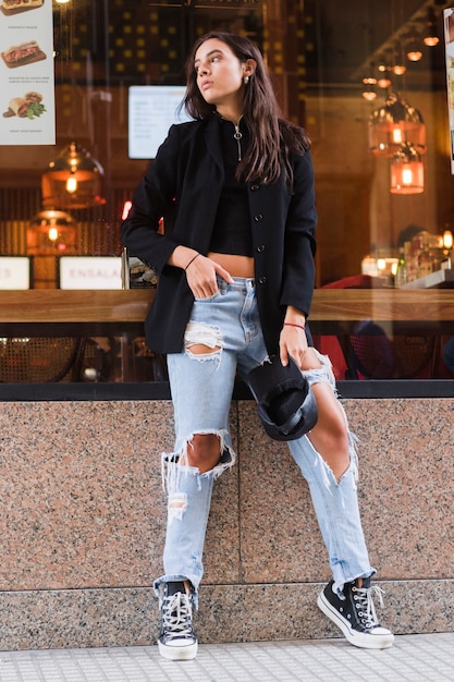 Free photo young woman leaning on glass of restaurant wearing ripped jeans holding black cap in hand