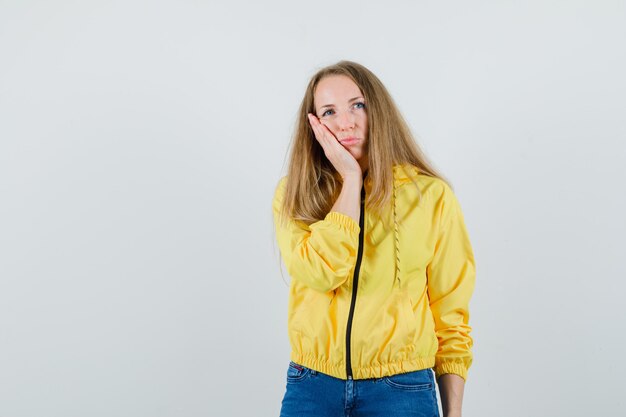 Young woman leaning hands on cheek in yellow bomber jacket and blue jean and looking regretful. front view.