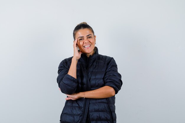 Young woman leaning head on hand in puffer jacket and looking cheerful. front view.