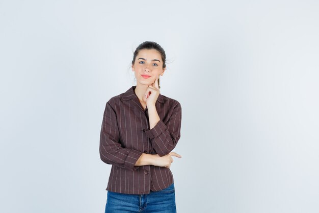 Free photo young woman leaning jowl on hand while posing in striped shirt, jeans and looking charming , front view.