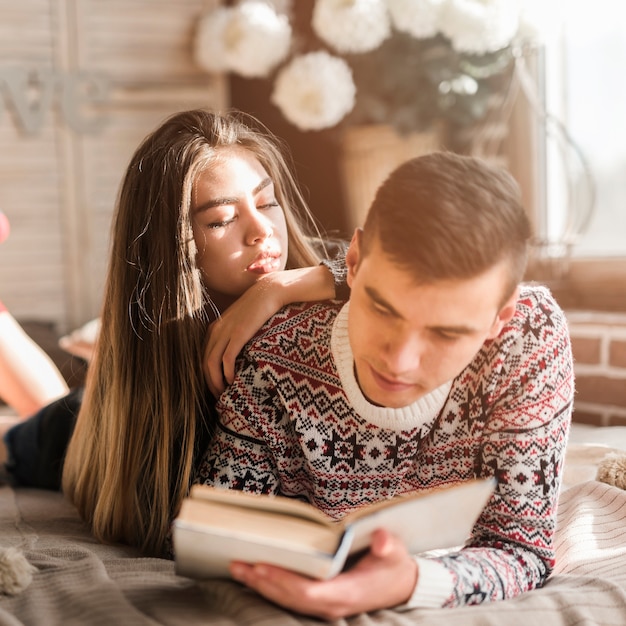 Young woman leaning on man's shoulder reading book on bed