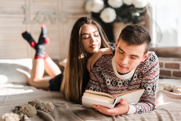 Young woman leaning on man's shoulder reading book on bed