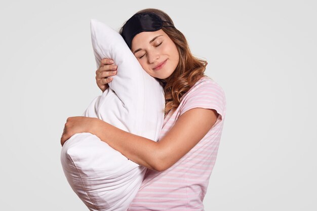 Young woman leans on pillow, wears pajamas and eye mask, stands against white, has sleepy expression