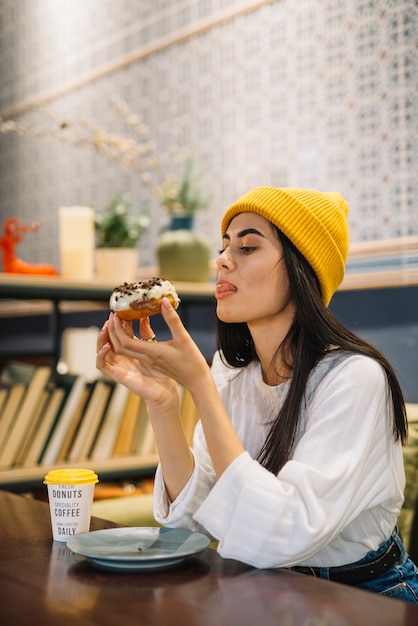 Free photo young woman licking lips and holding dessert near cup at table