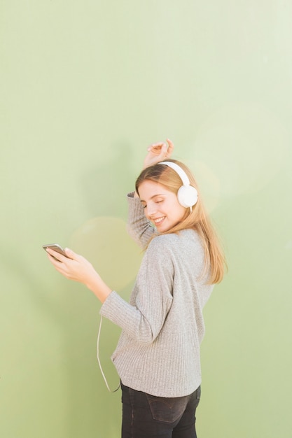 Young woman listening music on headphone through mobile phone dancing against mint green background