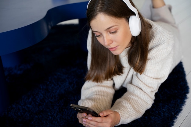 床に横たわっている間彼女のスマートフォンを使用して音楽を聴いている若い女性