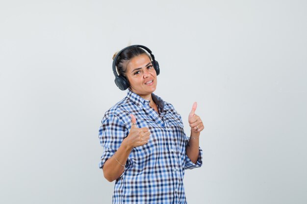 Young woman listening music while showing thumb up in checkered shirt and looking glad. front view.