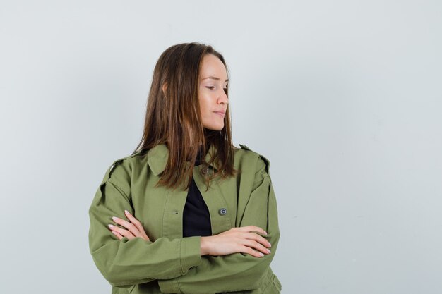 Young woman listening someone while standing with crossed arms in green jacket and looking careful , front view.