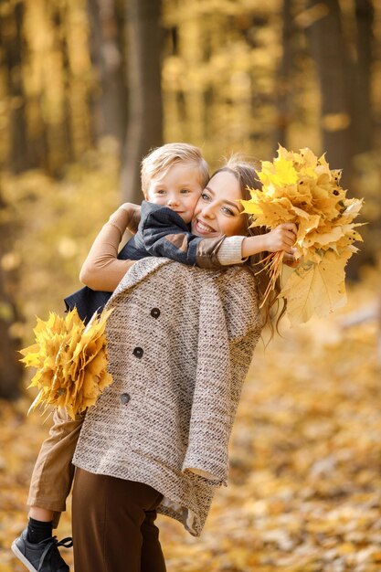 Молодая женщина и маленький мальчик в осеннем лесу. Женщина держит сына на руках. Мальчик в модной одежде и с желтыми листьями в руках.