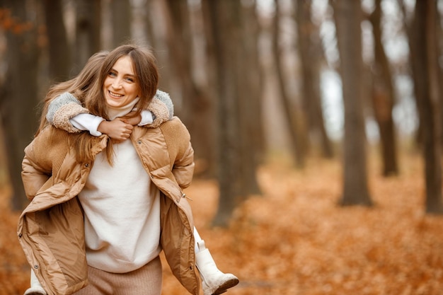 Giovane donna e bambina nella foresta di autunno donna portare la figlia sulle spalle ragazza che indossa un abito grigio moda con una giacca