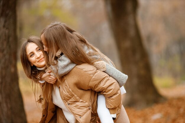 Young woman and little girl in autumn forest Woman carry her daughter piggyback Girl wearing fashion grey dress with a jacket