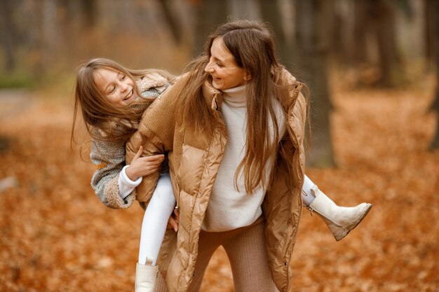 Young woman and little girl in autumn forest Woman carry her daughter piggyback Girl wearing fashion grey dress with a jacket