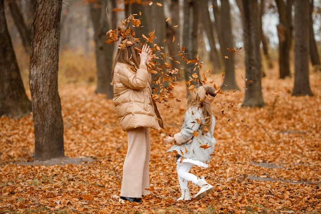 가을 숲의 젊은 여자와 어린 소녀 여자와 그녀의 딸이 마른 잎을 공중에 던지고 재킷과 함께 패션 회색 드레스를 입고 소녀