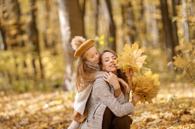 Young woman and little girl in autumn forest. Woman hugging her daughter. Girl wearing fashion clothes and holding yellow leaves.