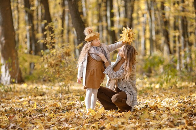 Young woman and little girl in autumn forest. Woman near her daughter who holding yellow leaves. Girl wearing fashion clothes.