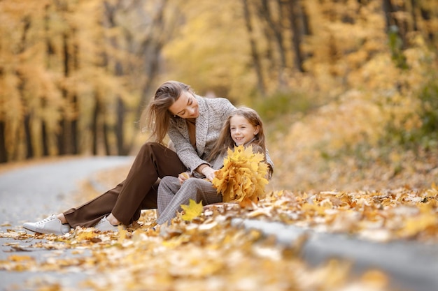 Young woman and little girl in autumn forest. Woman sitting near her daughter who holding yellow leaves. Mother hugging her daughter.