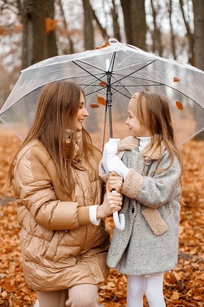 젊은 여자와 투명 우산 아래 가을 숲에 서 있는 어린 소녀 그녀의 어머니를 찾고 어린 소녀 패션 회색 재킷과 여자 갈색 하나를 입고 소녀