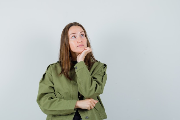 Young woman looking away while leaning her hand in green jacket and looking pensive. front view.
