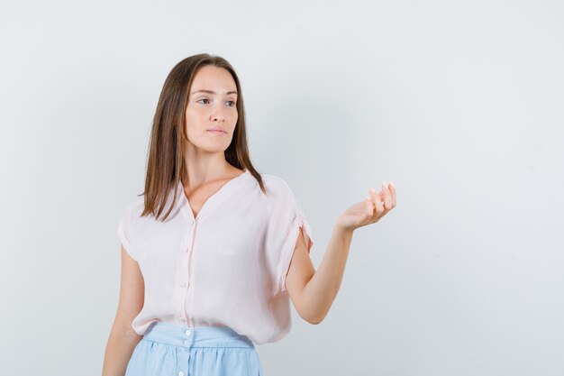 Young woman looking away with raised palm in t-shirt, skirt and looking pensive , front view.