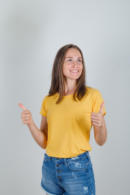 Молодая женщина смотрит в сторону с большими пальцами руки в желтой футболке, шортах и выглядит весело