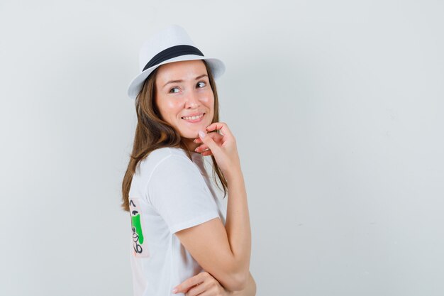 Молодая женщина, оглядываясь назад, думая в белой футболке, шляпе и выглядя оптимистично. .
