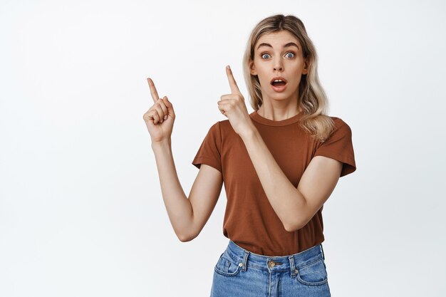 Young woman looking impressed, pointing fingers at upper left corner, showing shocking news, standing on white