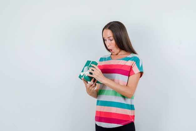 Young woman looking into gift box in t-shirt, pants and looking amazed , front view.