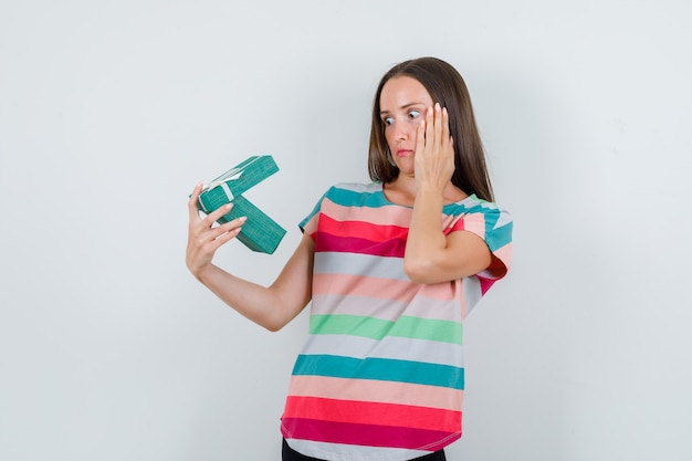 Young woman looking into gift box in t-shirt, pants and looking surprised , front view.