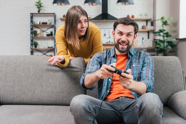 Young woman looking at man playing video game with console shrugging