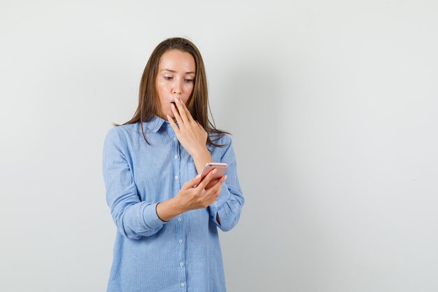 Young woman looking at mobile phone in blue shirt and looking shocked