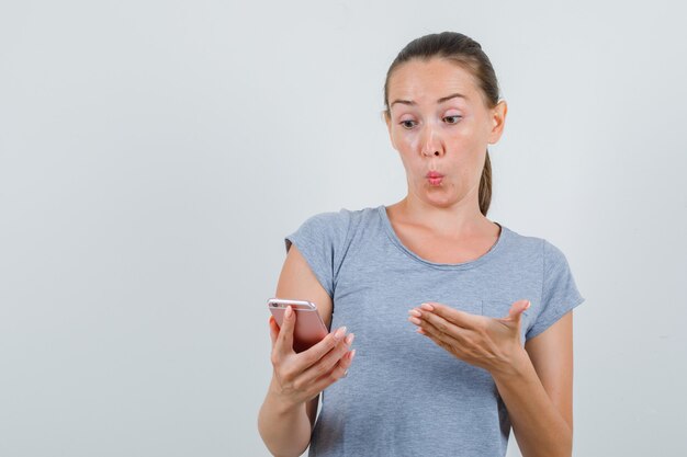Молодая женщина смотрит на мобильный телефон в серой футболке и выглядит удивленным. передний план.