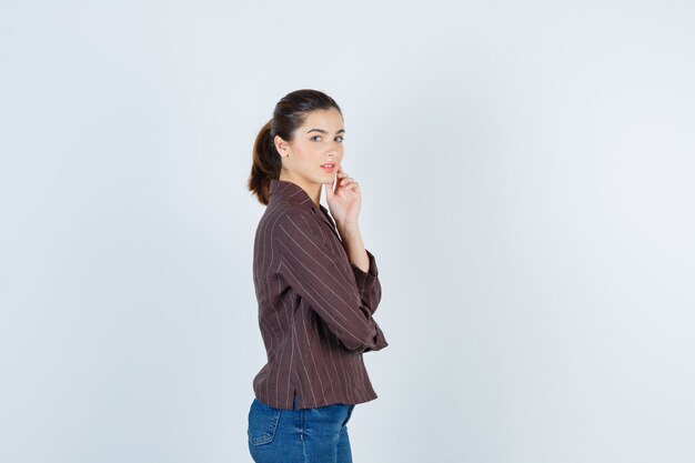 Young woman looking over shoulder, with hand on chin in striped shirt, jeans and looking charming. front view.