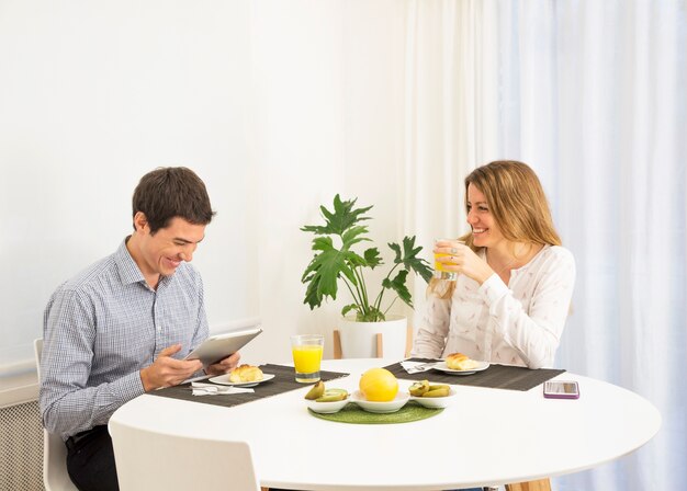 Молодая женщина, глядя на улыбающийся человек, используя цифровой планшет на завтрак стол