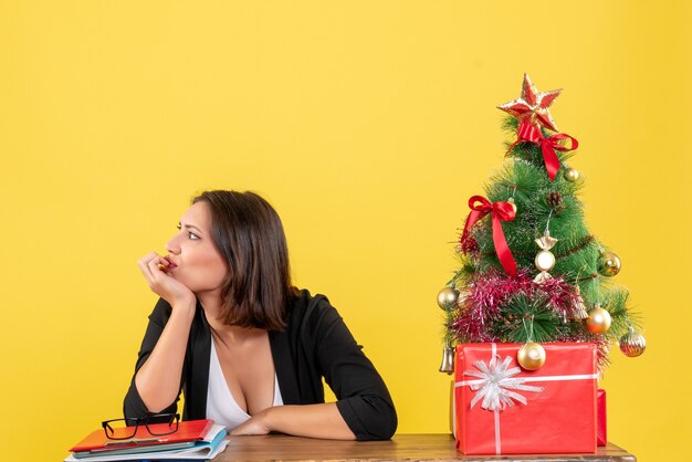 Young woman looking at something sitting at a table near decorated Christmas tree at office on yellow 