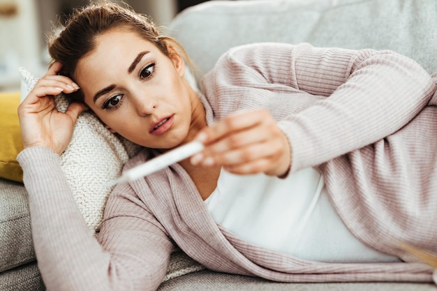 Young woman looking at thermometer in disbelief while measuring her body temperature at home