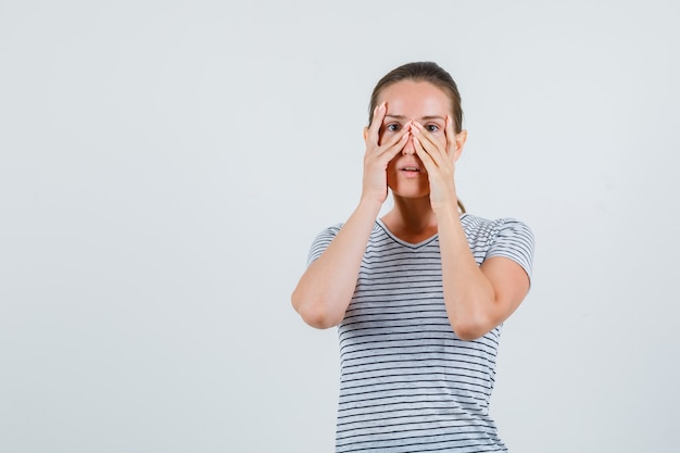 Young woman looking through fingers in striped t-shirt and looking scared , front view.