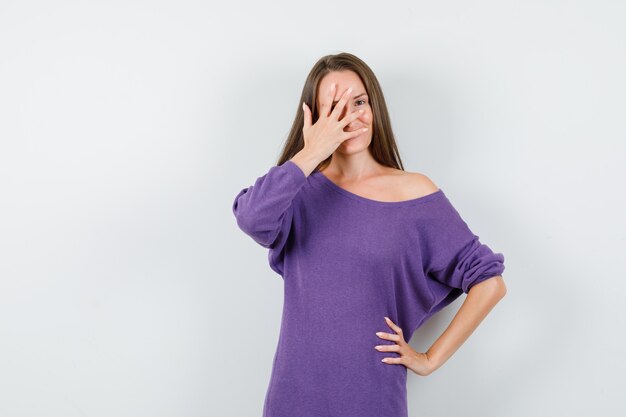 Young woman looking through fingers in violet shirt and looking positive , front view.