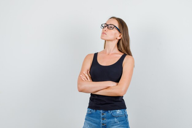 Young woman looking up with crossed arms in singlet, shorts, glasses and looking pensive