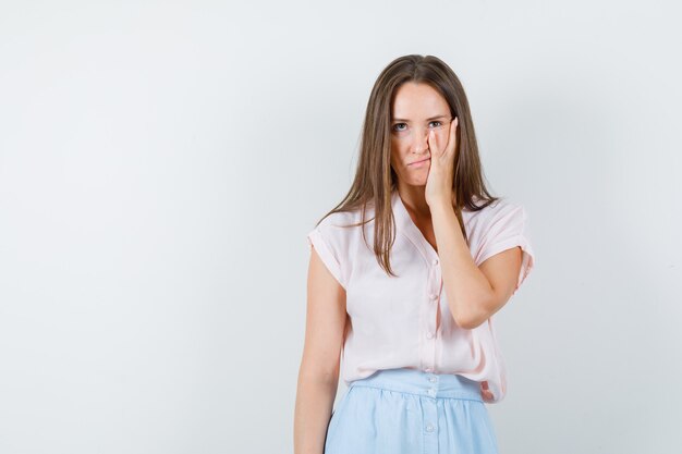 Young woman looking up with hand on cheek in t-shirt, skirt and looking nervous. front view.